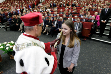 In the background, many people sitting in red chairs. In the foreground a young girl dressed elegantly, smiling, with her back to the camera an older man who is touching her shoulder with a rector's sceptre.