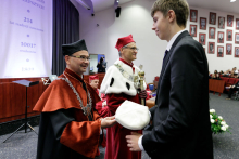 Wysoki młody mężczyzna ubrany w garnitur odbiera czapkę - symbol immatrykulacji od mężczyzny w średnim wieku ubranego w czarno-pomarańczową togę dziekańską.