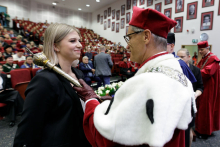 Na pierwszym planie, po lewej młoda dziewczyna z jasnymi włosami ubrana elegancko, po prawej mężczyzna w średnim wieku w todze czerwono-białej dotyka jej prawego ramienia berłem rektorskim. W tle wiele osób.