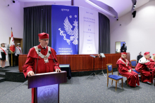 A man in a red academic toga with a chain around his neck stands behind the lectern and speaks into the microphone. Behind him, several people are seated in chairs and a banner post. In the background a decoration in blue and white.