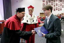 The young man - on the right receives a navy blue briefcase from the hands of the middle-aged man - on the left, dressed in a red and black academic toga. They shake hands. In the background stands a third man dressed in a red and white toga.