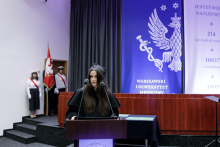 Młoda kobieta z ciemnymi włosami, ubrana w czarną togę akademicką stoi za mównicą. Za nią poczet sztandarowy oraz szara kotara i niebiesko-biała dekoracja.