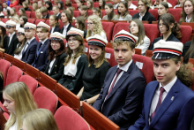 Grupa młodych osób ubranych elegancko, siedzą na czerwonych fotelach. Uśmiechają się do aparatu. Na głowie mają białe czapki z czerwonym otokiem i czarnym daszkiem.