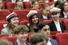 A group of young people dressed elegantly, sit in red chairs. They are smiling at the camera. On their heads they are wearing white caps with a red brim and a black visor.