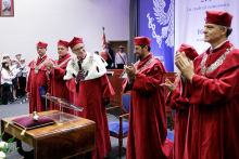 6 people dressed in red academic togas stand and applaud. Beside them is a table in brown and on it the rector's sceptre. Behind them a blue and white decoration.
