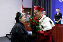 On the left, a middle-aged woman, wearing a black academic toga; on the right, a middle-aged man, wearing a white and red academic toga, with glasses, hands the woman a bouquet of flowers and shakes her hand.