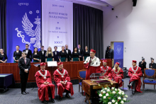 In the foreground, a group of people dressed in red togas. In the middle stands a middle-aged man and speaks into a microphone. In front of him is a table and a basket with white roses. Behind them stands a group of young people dressed in black. Behind them a curtain and a white and blue decoration with an eagle.