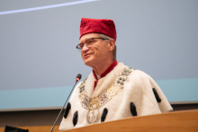 A middle-aged man, dressed in a red and white toga, with a red biretta on his head. He speaks into a microphone.