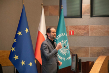 Mężczyzna w szarym garniturze stoi i przemawia do mikrofonu. Obok niego trzy flagi: UE, Polski i SGH