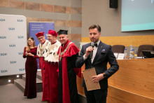 A group of people standing in the middle of the room. Some of them dressed in academic togas. The man on the right, dressed in a suit, holds a microphone in his hands and speaks.