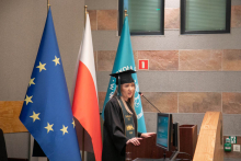 A young woman dressed in a black toga stands and speaks into a microphone. Next to her are three flags: EU, Poland and SGH.