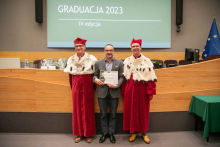 W tle ekran multimedialny, na środku auli stoi trzech mężczyzn, dwóch ubranych w czerwono-białe togi akademickie, mężczyzna w środku jest ubrany w garnitur, trzyma w rękach dyplom.