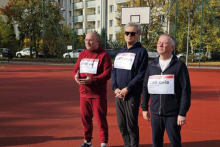 Trzech mężczyzn ubranych w stroje sportowe. Na bluzach mają przyklejone kartki z nazwiskami osób walczących o Ukrainę. Mężczyźni stoją na boisku szkolnym