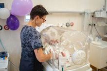 Kobieta ubrana w fartuch szpitalny stoi przy inkubatorze z niemowlęciem.