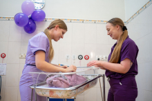Dwie kobiety ubrane w fioletowe fartuchy szpitalne stoją nad leżącym na łóżeczku noworodkiem.