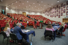 Aula uniwersytecka. Na widowni siedzi bardzo dużo osób. Z przodu na scenie trzy stoły, przy których siedzi kilka osób. przy dwó