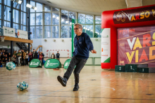 Mężczyzna w średnim wieku, ubrany na sportowo jest na środku sali gimnastycznej i kopie piłkę do nogi.
