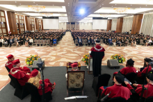 From the back, a group of people dressed in academic togs sit on chairs on a raised platform, in front of them a crowd of people sitting on chairs, dressed in black academic togs.