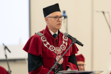 A middle-aged man, wearing glasses and dressed in a red and black toga speaks into a microphone.