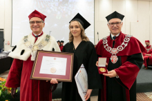 Three people, on the left and on the right men, in the middle a young woman who holds a framed diploma in her hands. They stand and smile for the camera. They are dressed in academic togas.