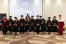 A group of young people dressed in black academic togas stand with diplomas in their hands, smiling at the camera. Next to them is the rector dressed in a red and white academic toga.