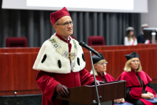 Na pierwszym planie mężczyzna ubrany w biało-czerwoną togę akademicką. Stoi przy mównicy i przemawia. Po jego lewej stronie siedzi kilka osób. W tle ekran multimedialny. 