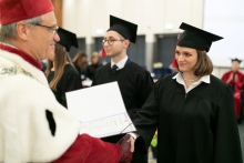 A group of people dressed in black academic togas and a man dressed in a white-red toga handing one person an open-covered diploma.