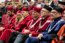 Grupa osób w togach akademickich w różnych kolorach. Siedzą na czerwonych fotelach i patrzą przed siebie.
