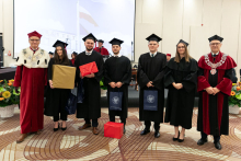 A group of young people dressed in black academic togas stand with diplomas in their hands, smiling at the camera. Next to them is the rector dressed in a red and white academic toga and the dean dressed in a black and red tog