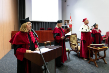 Na pierwszym planie kobieta w czerwono-czarnej todze. Stoi za mównicą i przemawia. Po jej lewej stronie, nieco z tyłu stoi kilka osób ubranych w togi akademickie. 