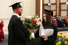 A young man dressed in a black academic toga hands a toga-clad young woman a bouquet of flowers. They smile.