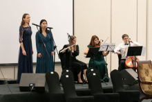 A few people on a raised platform, two women stand at microphones and sing, three people sit and play instruments.