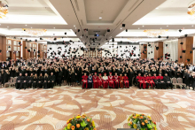 A group of people, graduates - dressed in black togas - stand and throw birettas. In front of them sits a group of people dressed in academic attire.