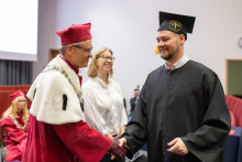 Na zdjęciu trzy osoby: w środku młoda kobieta, na przedzie dwóch mężczyzn ubranych w togi: rektor i absolwent. Ściskają sobie dłonie i uśmiechają się.