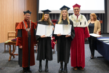 Na zdjęciu cztery osoby: w środku dwie młode kobiety, trzyma w rękach dyplom. Po jej obu stronach stoją mężczyźni - rektor i dziekan.