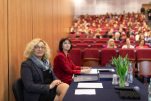 Na pierwszym planie dwie elegancko ubrane kobiety, patrzą w bok. W tle grupa osób, uczestników konferencji, siedzą i patrzą przed siebie.