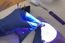 Ręka w niebieskiej rękawiczce, która trzyma narzędzie dentystyczne. Ręka pracuje nad czymś oświetlonym jasnym światłem. W tle znajdują się różne narzędzia dentystyczne, w tym kilka kolorowych końcówek i strzykawka.