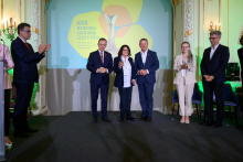 Na zdjęciu znajduje się 6 osób stojących na scenie. W tle widać ekran z napisem "XXII NAGRODA ZAUFANIA ZŁOTY OTIS". Osoby są elegancko ubrane, niektóre z nich trzymają nagrody lub dyplomy. Po bokach sceny znajdują się dekoracje i oświetlenie. W centralnym punkcie znajduje się nagrodzona prof. Lidia Rudnicka. Obok niej stoi dwóch mężczyzn, wszyscy pozują do zdjęcia. Prof. Rudnicka trzyma w uniesionej ręce nagrodę. Po bokach sceny znajdują się dekoracje i oświetlenie.