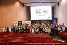 A large group of people standing on a stage in the auditorium. In the background is a screen with the words “20th WIMC Closing Ceremony.” Banners and posters related to the event are also visible on the stage. In front of the stage are rows of red chairs.