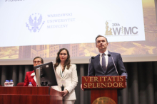 Młode kobiety Maria Żmijewska i Emilia Włoszek oraz mężczyzna - Mariusz Tomaniak. Wszyscy są elegancko ubrani. Mężczyzna stoi za mównicą i przemawia. Za nimi znajduje się duży ekran wyświetlający logotypy "Warszawski Uniwersytet Medyczny" oraz "20th WIMC". Tło ekranu przedstawia zdjęcie budynków.