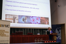 The speaker, Prof. Anita Arsovska, standing in the auditorium, stands behind the lectern and shows a presentation. Behind her, on a large multimedia screen, a slide with captions and photos is displayed. Next to it, on the left, a rollup of the congress with the WIMC caption and logo can be seen.