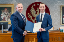 Dwóch mężczyzn, elegancko ubranych, pozuje do zdjęcia, uśmiechają się. Mężczyzna po prawej trzyma w rękach teczkę.