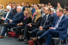 Grupa ludzi siedzących w sali konferencyjnej. Wszyscy są elegancko ubrani. W tle widać więcej osób, które również uczestniczą w wydarzeniu.