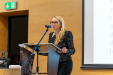 Elegancko ubrana kobieta z jasnymi włosami i w okularach, prof. Agnieszka Cudnoch-Jędrzejewska. Stoi za mównicą i przemawia. Za nią ekran z testem przemówienia po angielsku. Widać też drewniane ściany sali.