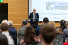 Mężczyzna w garniturze dr Artur Drobniak. Stoi za mównicą i przemawia do publiczności. W tle widoczny jest ekran z prezentacją, na którym znajdują się teksty i obrazy. Publiczność siedzi na krzesłach.