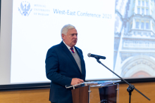 Pierwszy prelegent konferencji prof. Mark S. Wolff z Uniwersytetu w Pensylwanii. Starszy mężczyzna z siwymi włosami, w garniturze. Stoi przy mównicy z mikrofonem. W tle widać ekran z napisem "MEDICAL UNIVERSITY OF WARSAW" oraz "West-East Conference 2025".