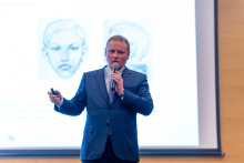 Mężczyzna w średnim wieku, elegancko ubrany, trzyma w jednej ręce mikrofon, drugą gestykuluje. W tle znajduje się ekran projekcyjny, na którym wyświetlane są dwa obrazy przedstawiające ludzkie twarze.