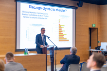 Jeden z prelegentów konferencji. Elegancko ubrany mężczyzna w okularach. Stoi przy mównicy i przemawia. Za nim znajduje się ekran z wyświetlonym slajdem zatytułowanym "Dlaczego otyłość to choroba?". Na ekranie widoczne są wykresy i tekst dotyczący tematu prezentacji. W tle widać kilka osób siedzących na krzesłach, słuchających przemówienia.