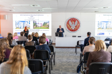 Na pierwszym planie znajduje się grupa ludzi, znajdująca się w sali konferencyjnej. Siedzą na krzesłach i słuchają prezentacji. Na scenie stoi dwóch prelegentów: mężczyzna przy mównicy oraz kobieta obok ekranu projekcyjnego. Na ekranie wyświetlane są slajdy z informacjami o konferencji.