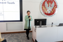 Prelegentka Dnia Endodontycznego. Elegancko ubrana kobieta, w zielonej marynarce, stoi przed ekranem multimedialnym, w dłoni trzyma mikrofon, uśmiecha się. Stoi obok biura z monitorem komputerowym. Na ścianie za nią widnieje duże godło WUM: biały orzeł na czerwonej tarczy, trzymający w szponach berło i laskę Eskulapa.